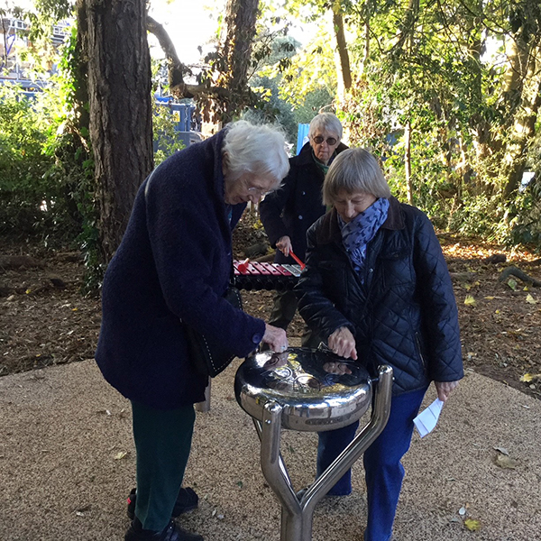 two ladies playing a round stainless steel tongue drum in the Aldeburgh Hopsital sensory garden