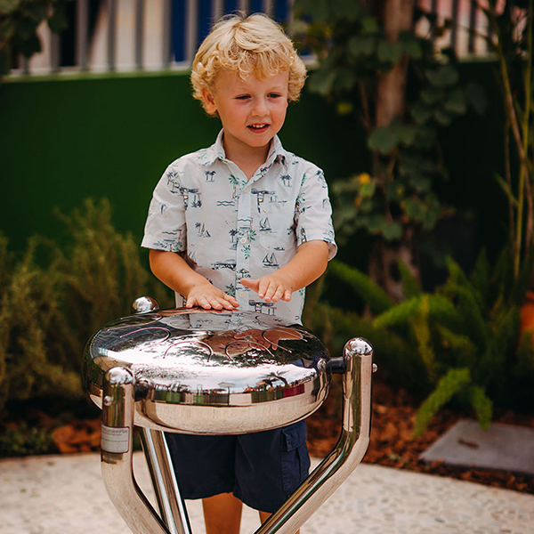 Small blonde boy playing an outdoor stainless steel tongue drum in a music park