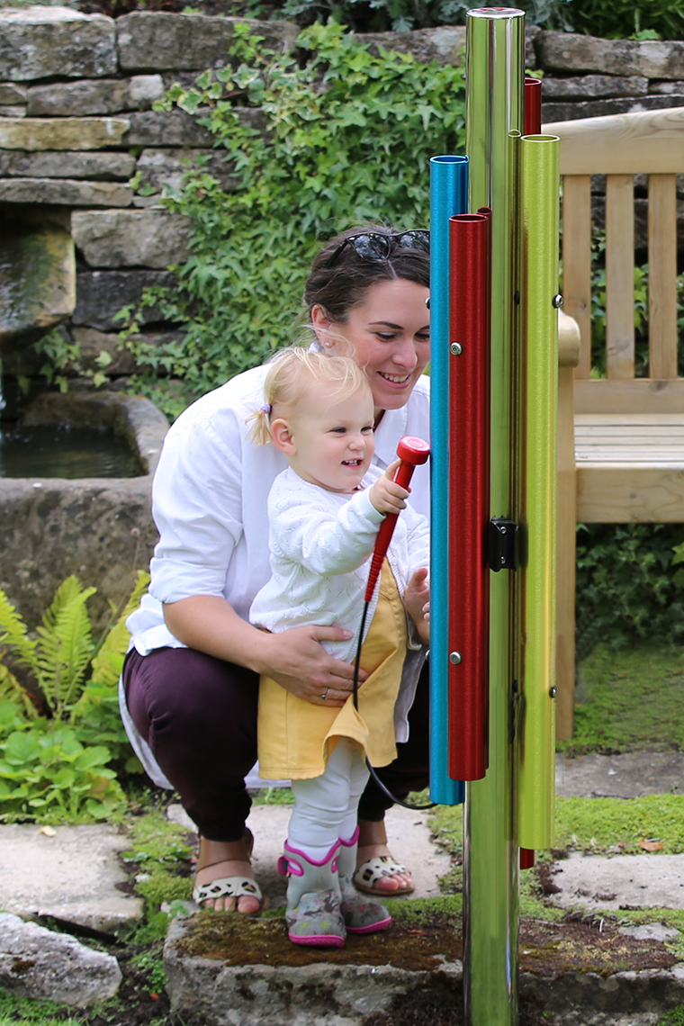 mother and child playing an outdoor musical instrument with four colorful chimes and red mallet