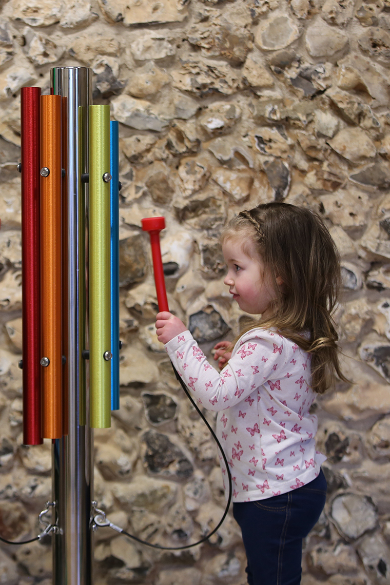 A little girl playing an outdoor musical instrument made of a single stainless steel post and five bright coloured chimes attached