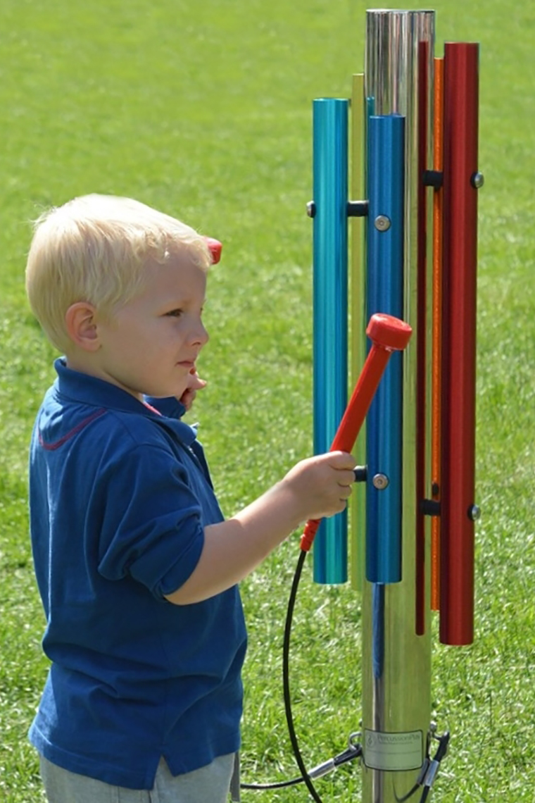 A little blonde boy playing on an outdoor musical instrument of five rainbow coloured chimes on a stainless steel post