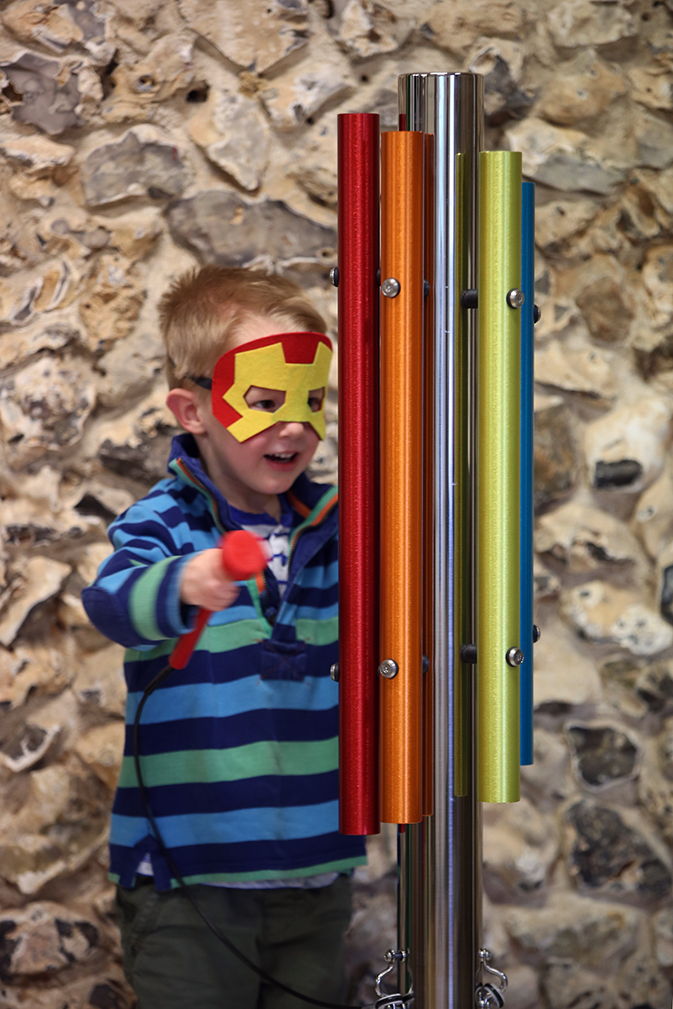 A little boy dressed as iron man playing an outdoor musical instrument made of a single stainless steel post and five bright coloured chimes attached
