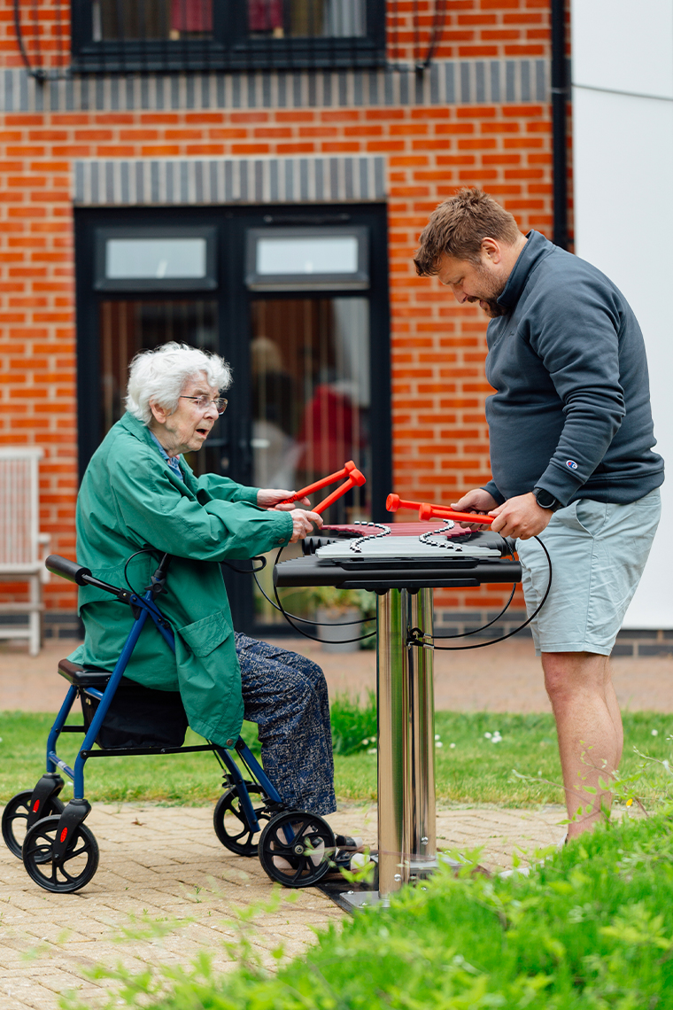 senior lady playing an outdoor musical instrument while seated in a wheelchair with a male carer