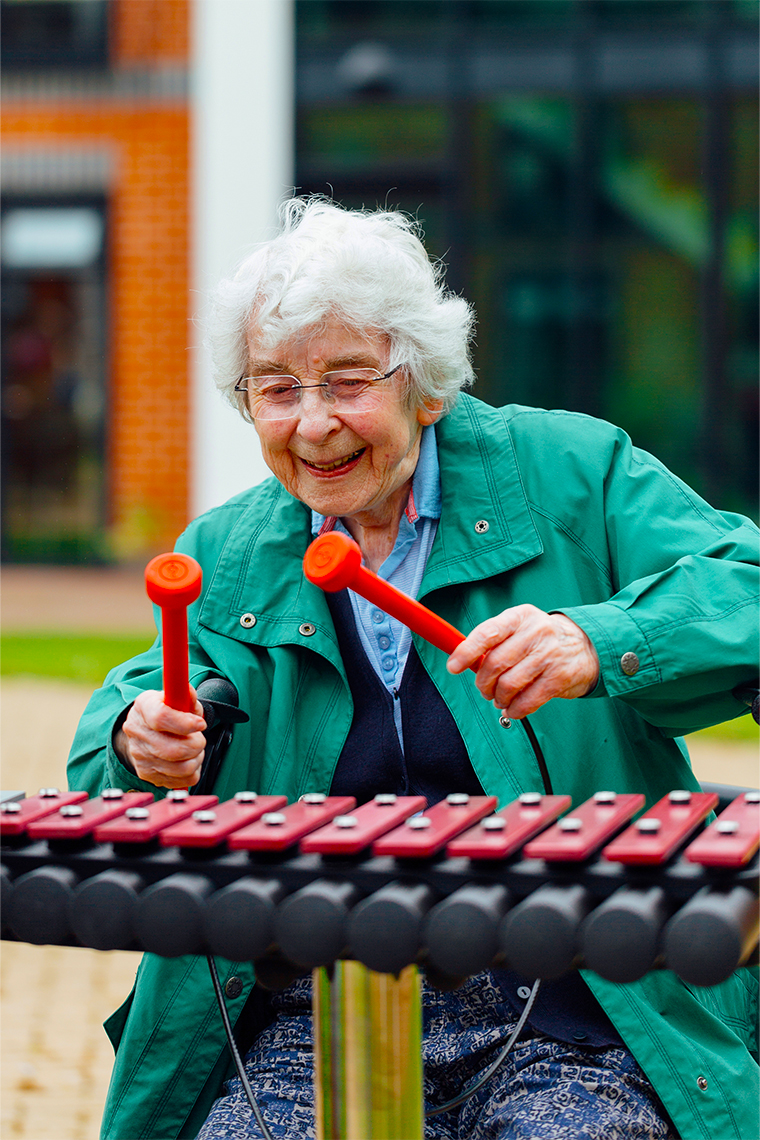 senior lady smiling and  seated playing an outdoor xylophone using red mallets